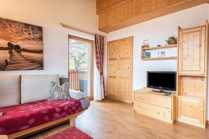 A room with a wooden floor, a sofa with patterned cushions, a TV on a wooden stand, and a painting of a sunset over a bridge.