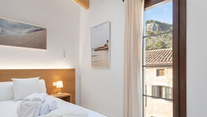 A hotel room with a bed, a nightstand with a lamp, a painting of a beach, and a window with a view of buildings. - Placeta Vella Turismo de Interior (Selva)