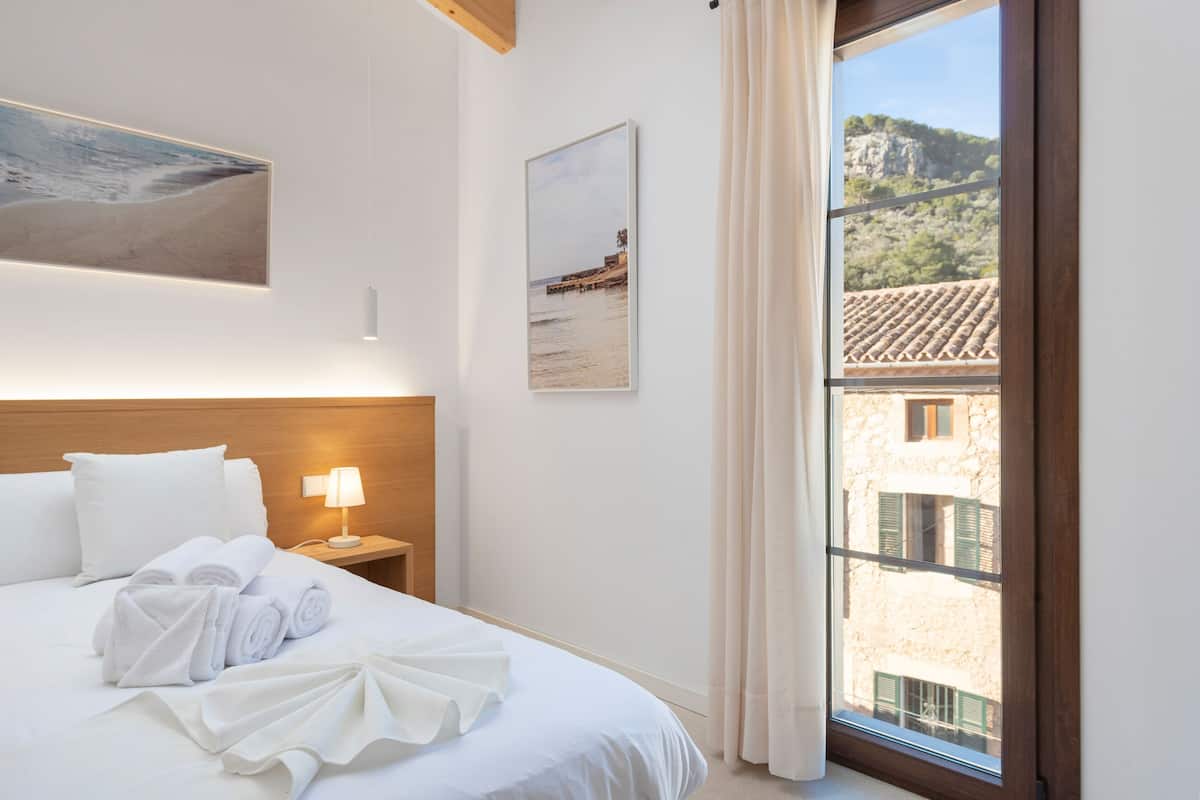 A hotel room with a bed, a nightstand with a lamp, a window with a view of buildings, and two framed pictures on the wall.