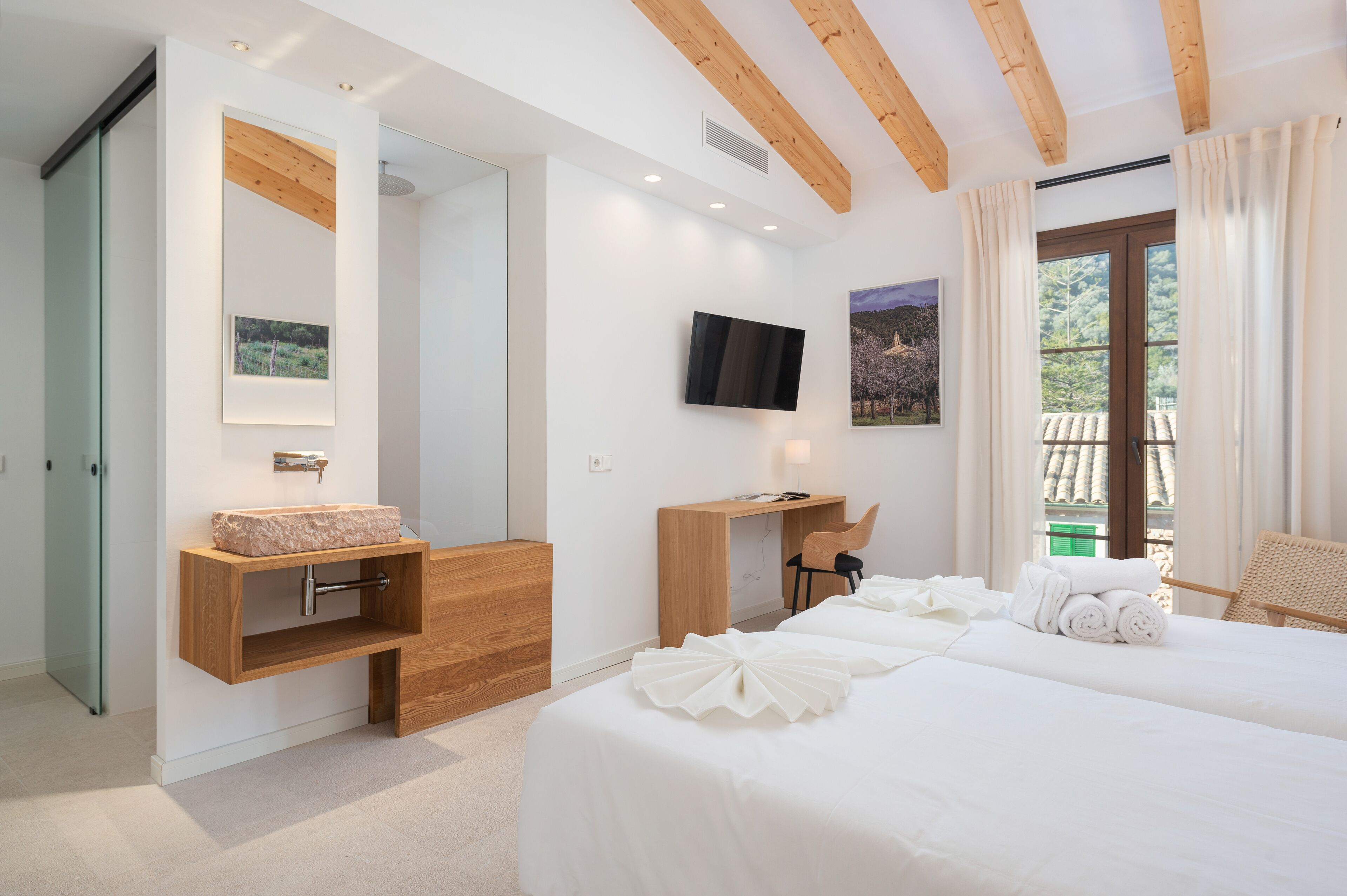 A modern bedroom with a wooden ceiling, a bed with white linens, a wooden desk, a television, and a large window with curtains.