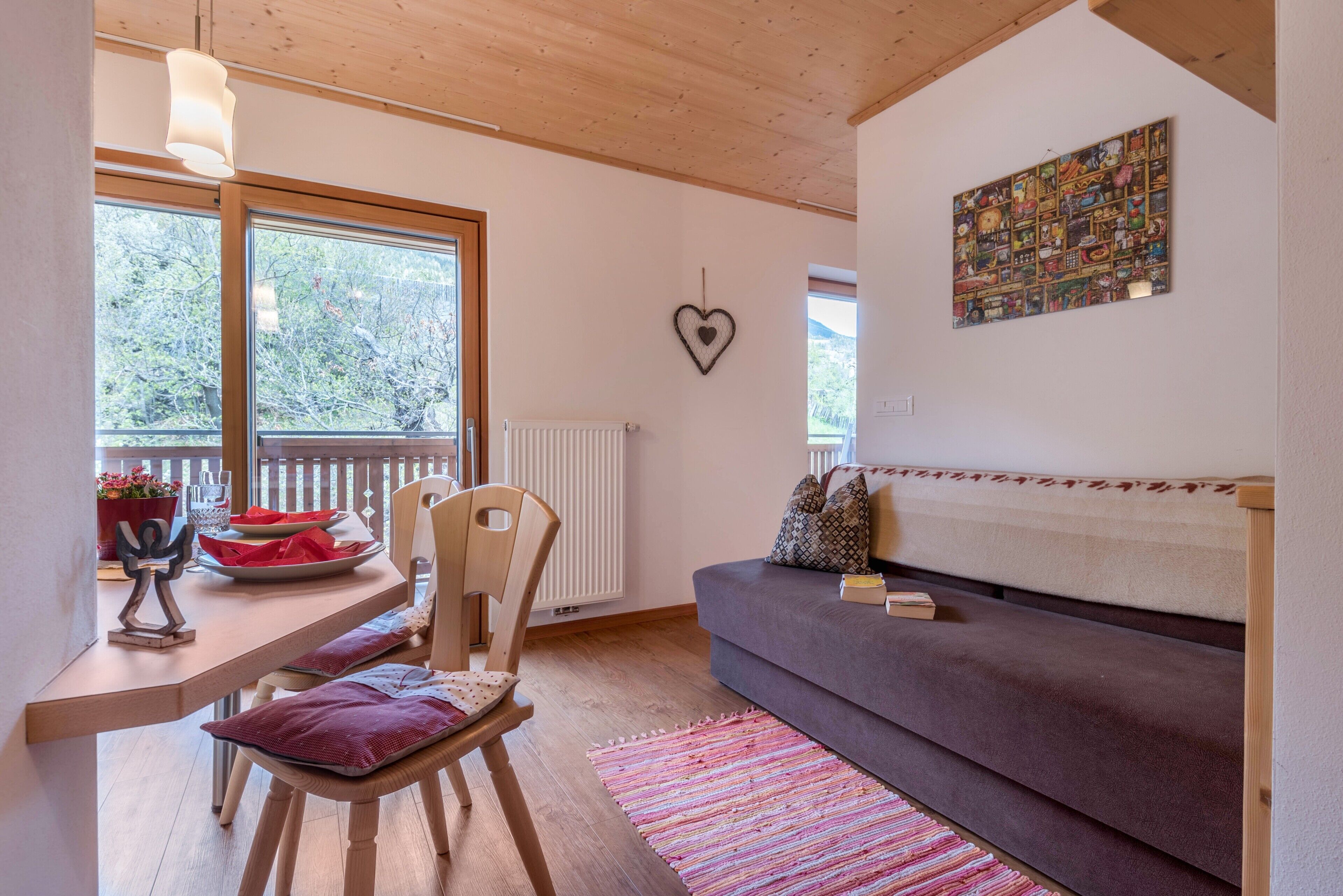 A cozy living area with a wooden ceiling, a sofa, a dining table with chairs, and a large window.
