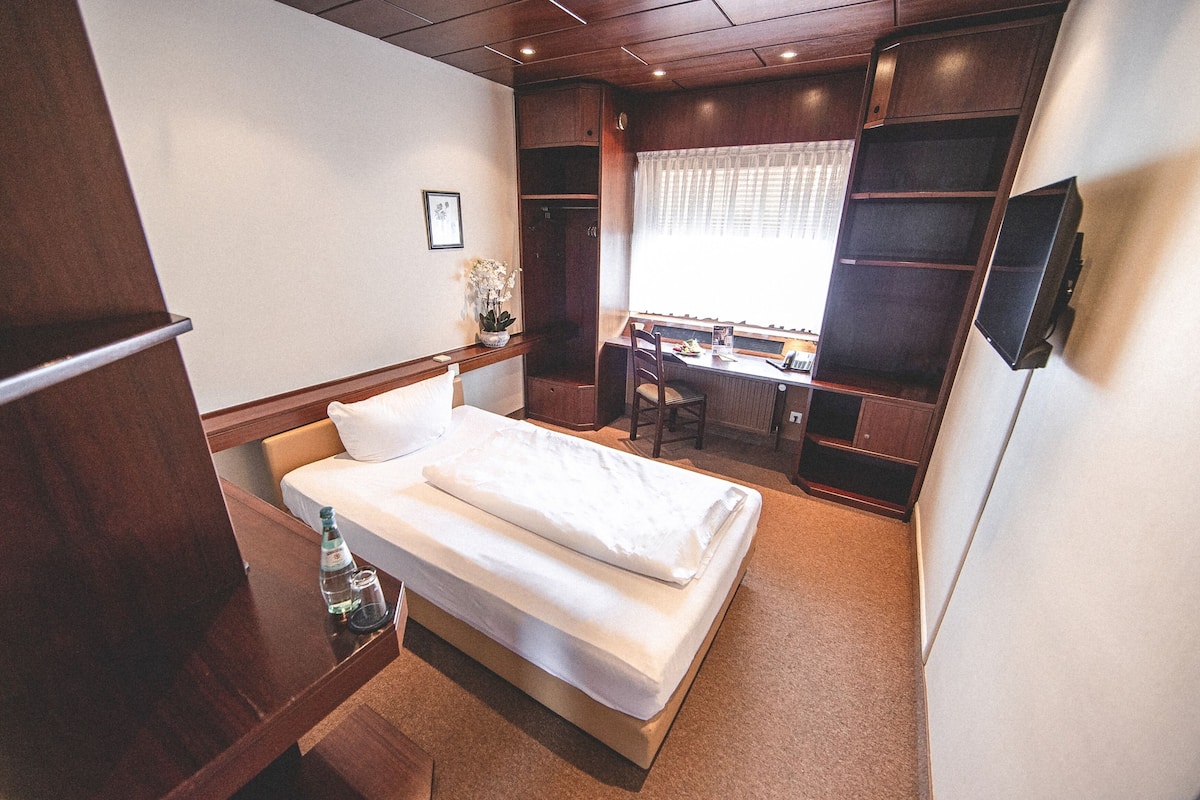 Habitación de hotel pequeña con cama, un escritorio, una silla y un ventanal con cortinas.
