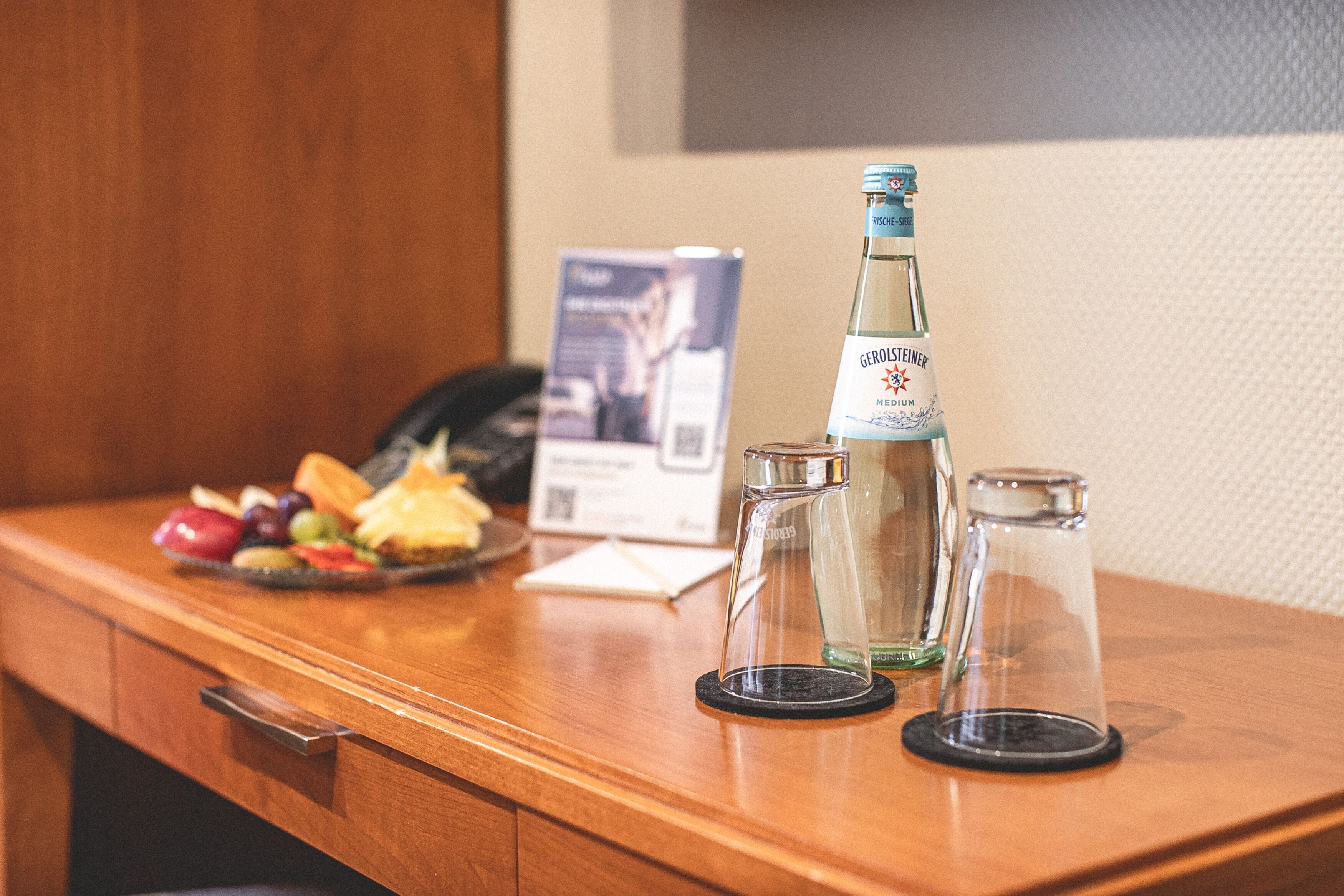 Ein Holztisch mit einer Gerolsteiner-Flasche, zwei Gläsern und einer Platte mit Obst und Käse.