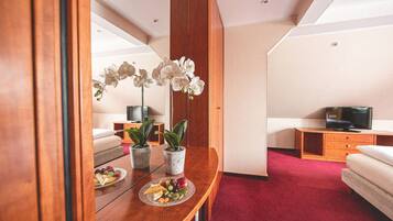 Ein Hotelzimmer mit einem Holztisch, zwei Topfpflanzen, einer Obstplatte, einem Bett, einem Fernseher und einem roten Teppich.