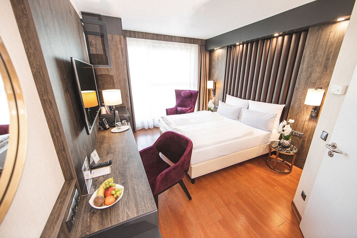 Een hotelkamer met een bed, een bureau met een schaal fruit, een stoel en een kleine tafel met een lamp.
