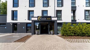 Un edificio moderno con un letrero que dice "Plaza Inn". Tiene un acceso adoquinado de ladrillo, plantas en macetas y un seto.