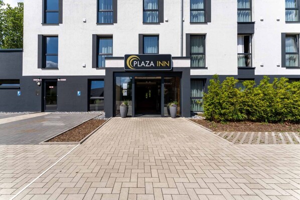 A modern building with a sign reading "Plaza Inn." It features a brick-paved entrance, potted plants, and a hedge. - PLAZA INN Recklinghausen (Recklinghausen)