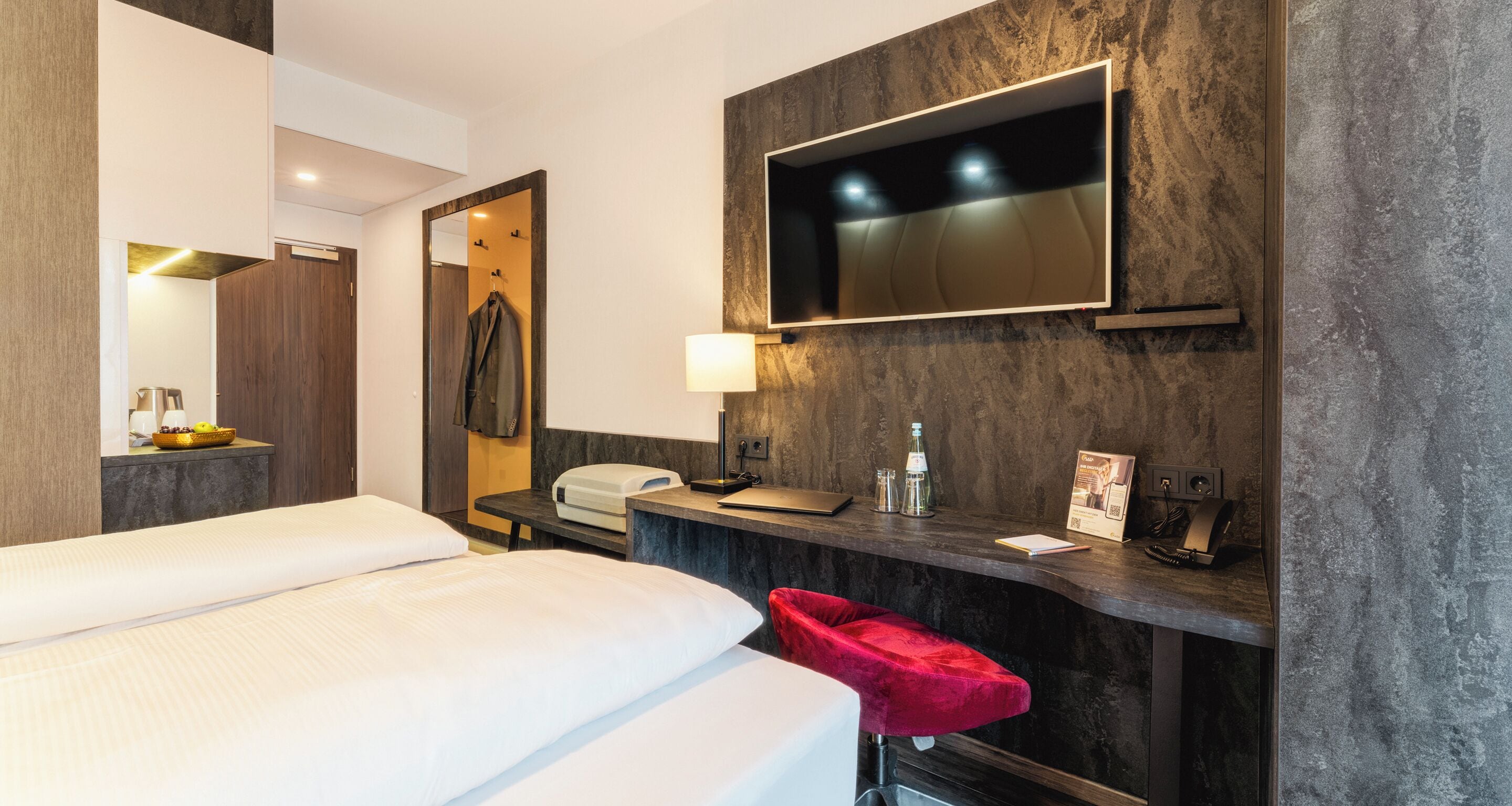 A hotel room with a bed, a desk with a lamp, a mirror, a TV mounted on the wall, and a red chair.