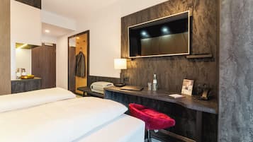 A hotel room with a bed, a desk with a lamp, a mirror, a TV mounted on the wall, and a red chair.
