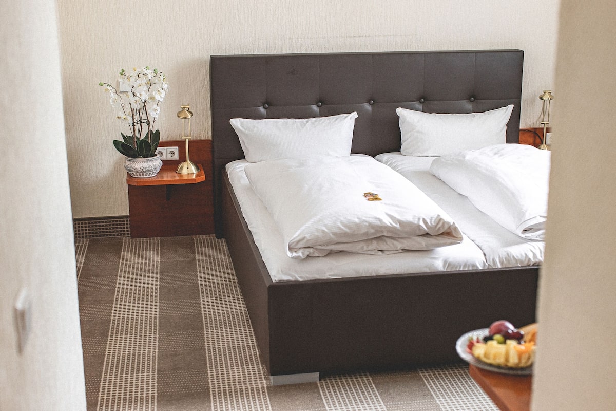 Una cama bien hecha con sábanas y almohadas blancas, una mesita de noche con lámpara y una pequeña planta en maceta.