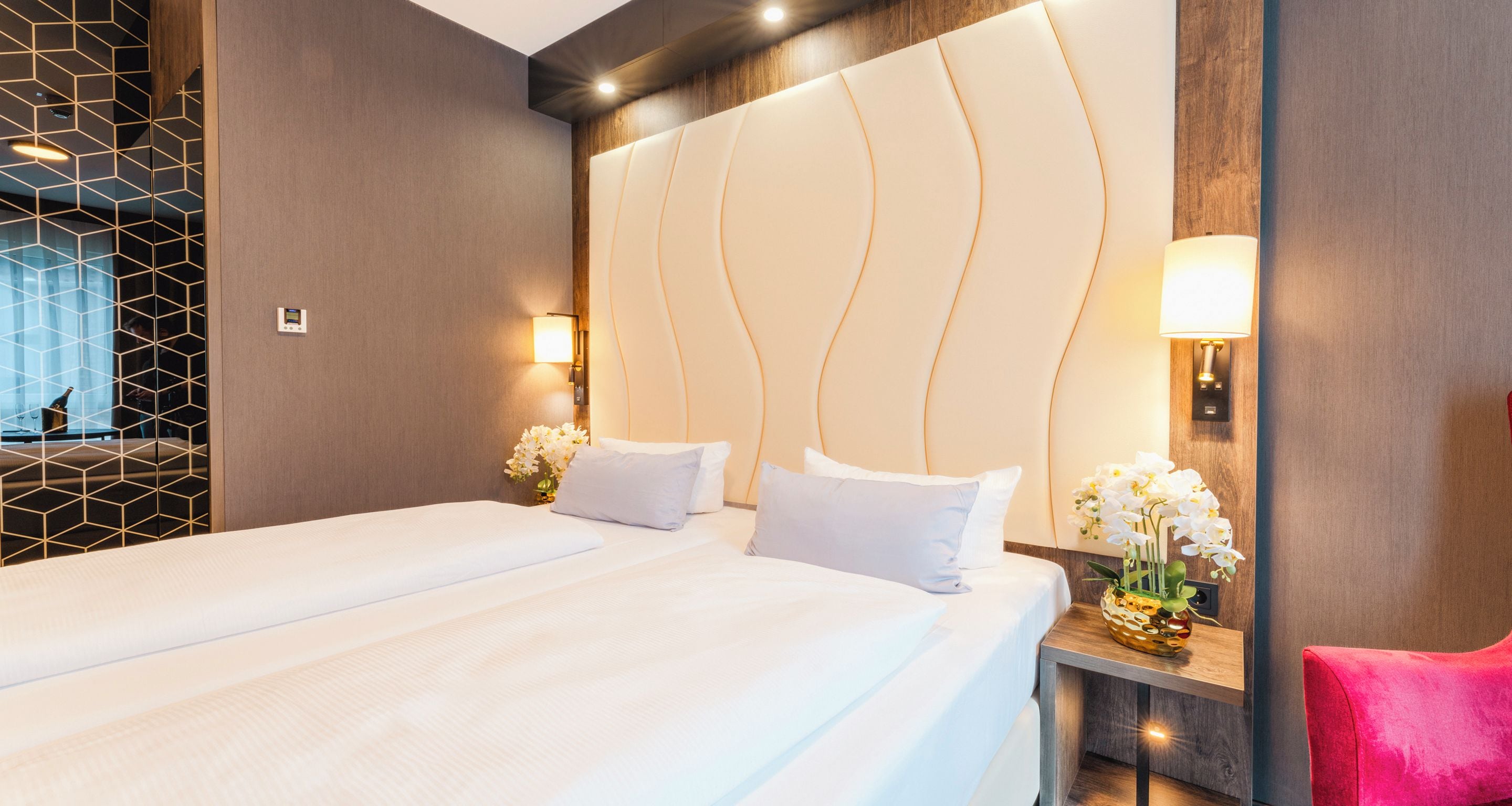 A hotel room with a large bed, two bedside tables, a nightstand, a red chair, and a decorative wall panel.