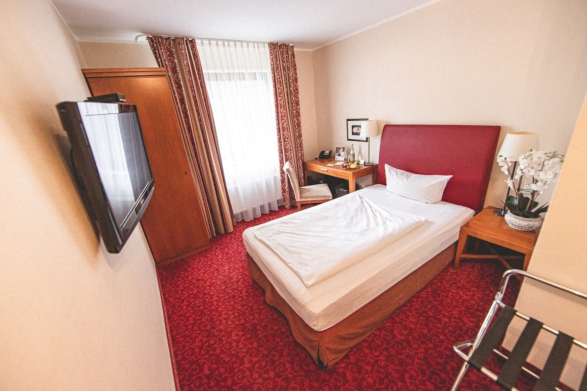 Habitación de hotel con cama, escritorio con lámpara, silla, televisión y ventana con cortinas.