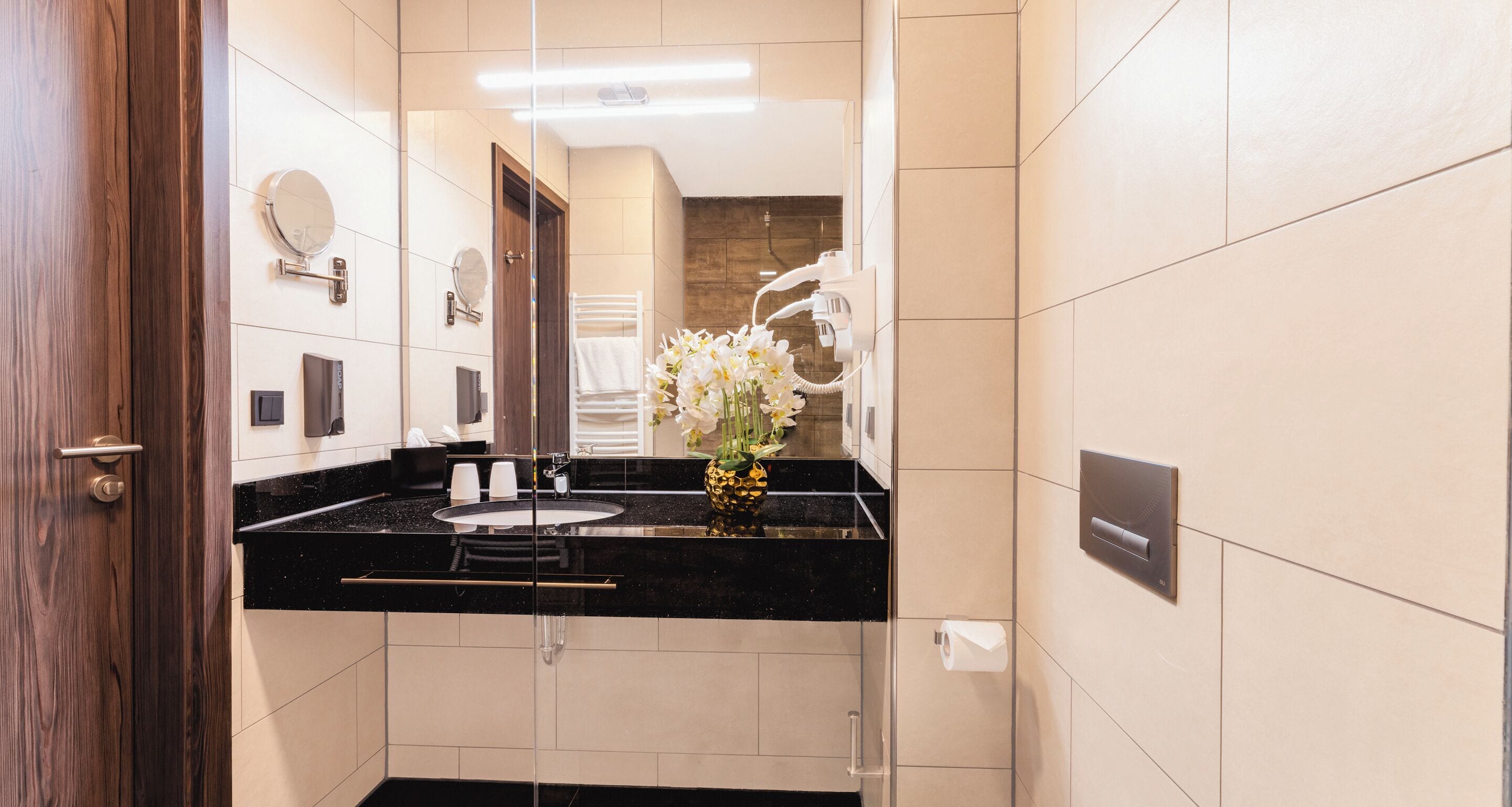 Un baño moderno con encimera negra, un espejo, toallero y un florero con flores blancas.