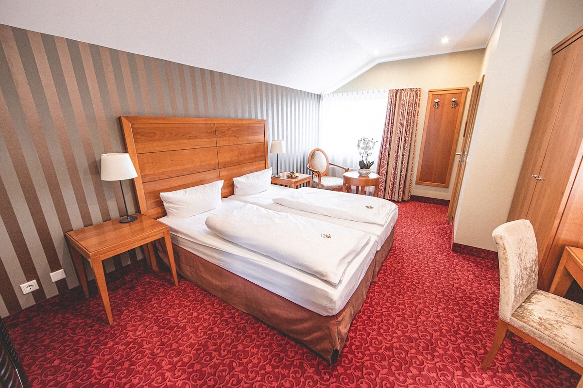 Una habitación de hotel con una cama grande, un cabecero de madera, una mesita de noche con una lámpara, una pequeña mesa con una silla, un espejo y un armario.