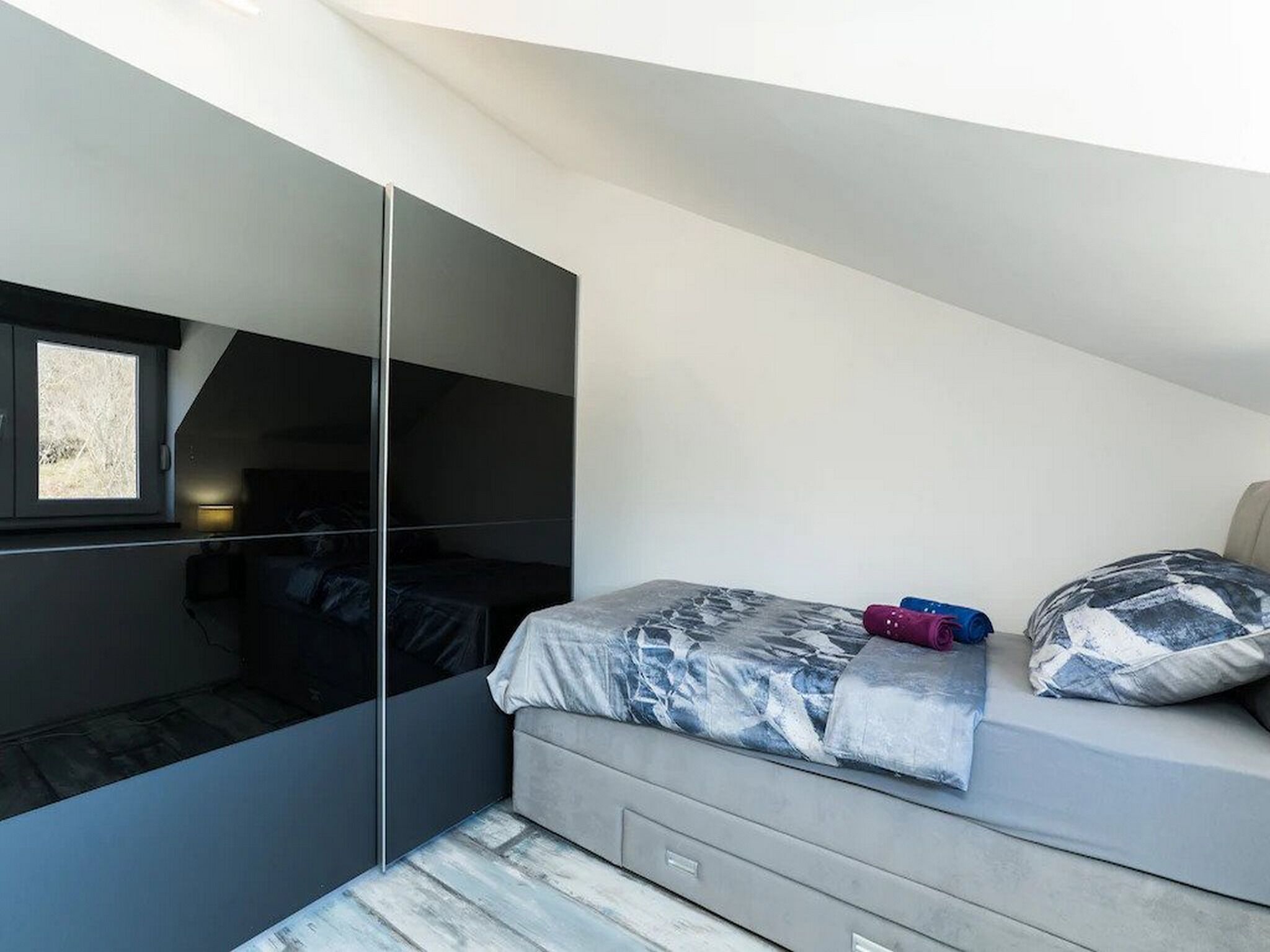 A modern bedroom with a large mirror, a bed with patterned bedding, and a window with a view.