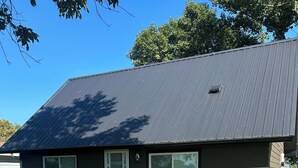 Une maison de plain-pied à l’extérieur brun foncé, avec un toit gris et un petit jardin devant, composé de pelouse et de quelques arbres.