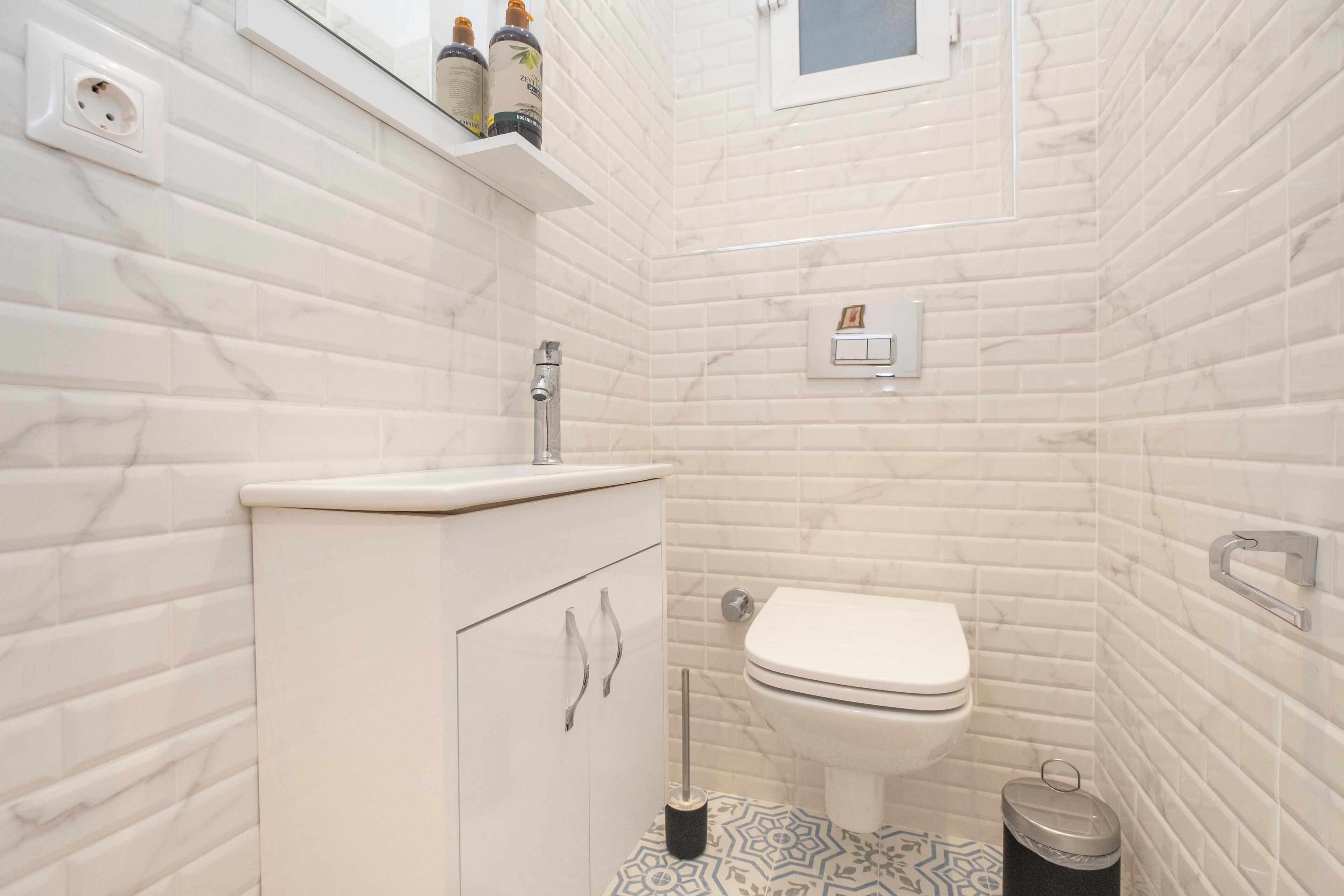 A bathroom with white marble tiles, a white sink, a toilet, and a trash bin.