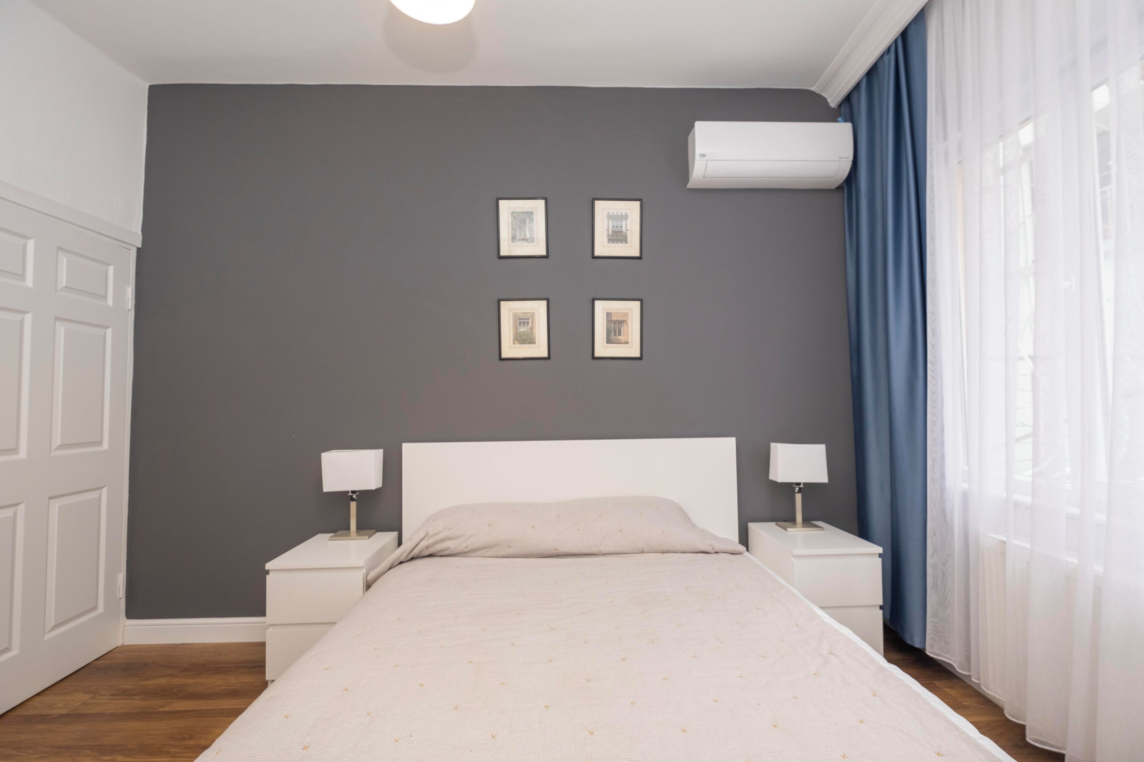 A bedroom with a bed, two bedside tables with lamps, a wall-mounted air conditioner, and four framed pictures on the wall.