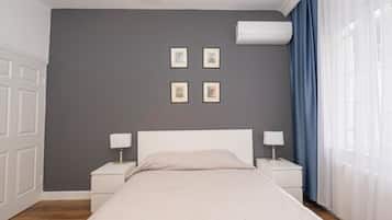 A bedroom with a bed, two bedside tables with lamps, a wall-mounted air conditioner, and four framed pictures on the wall.