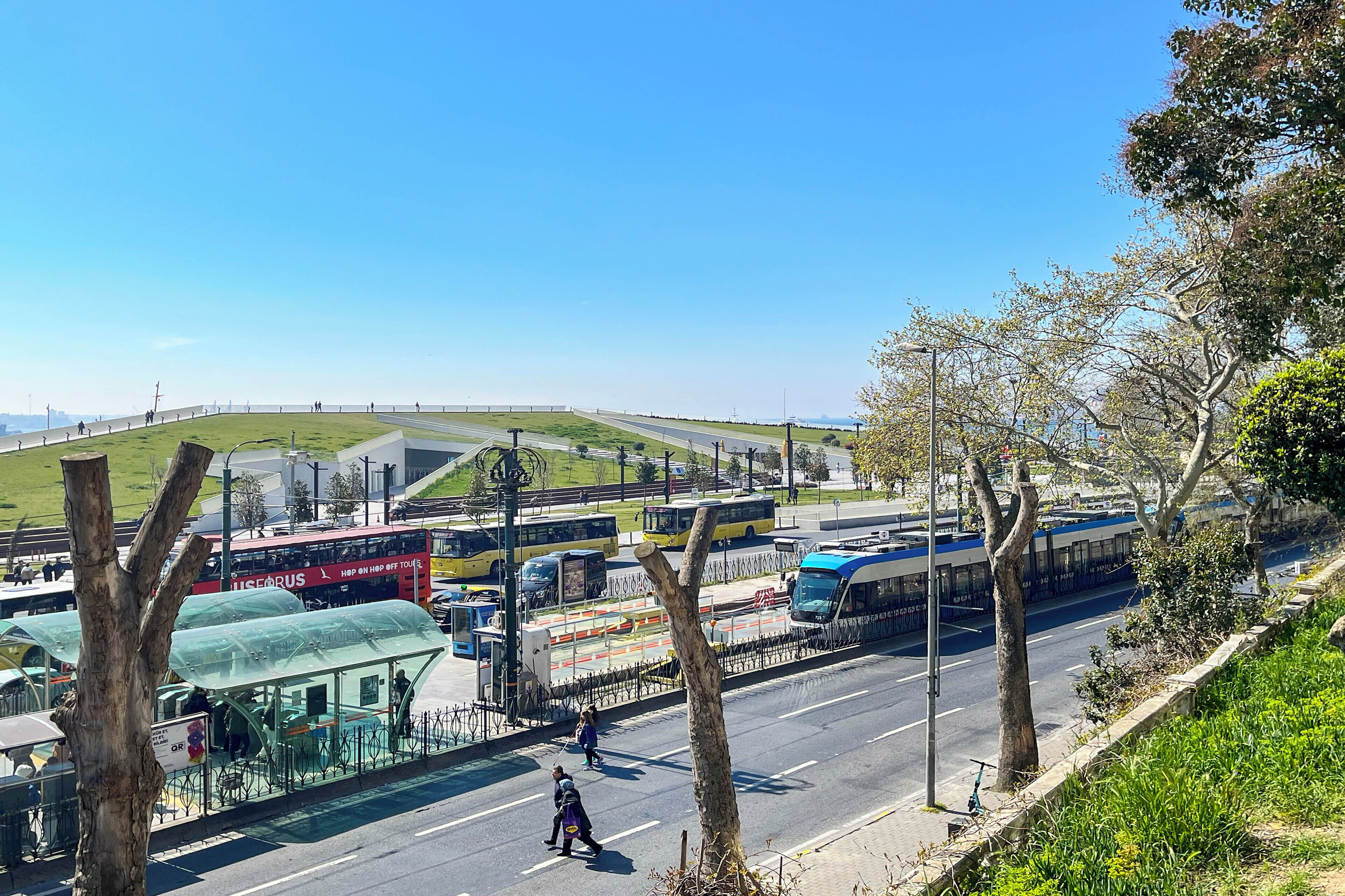 En busstation med flera bussar, ett övergångsställe och en gräsklädd kulle i bakgrunden.