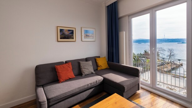 Een moderne woonkamer met een grijze modulaire bank, een houten salontafel en een groot raam met uitzicht op een waterpartij en een verre skyline.