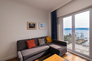 A modern living room with a grey sectional sofa, a wooden coffee table, and a large window offering a view of a body of water and a distant cityscape. - Trendy 2BR Apartment · Bosphorus & Dolmabahçe View · Central Beyoğlu (Beyoglu)