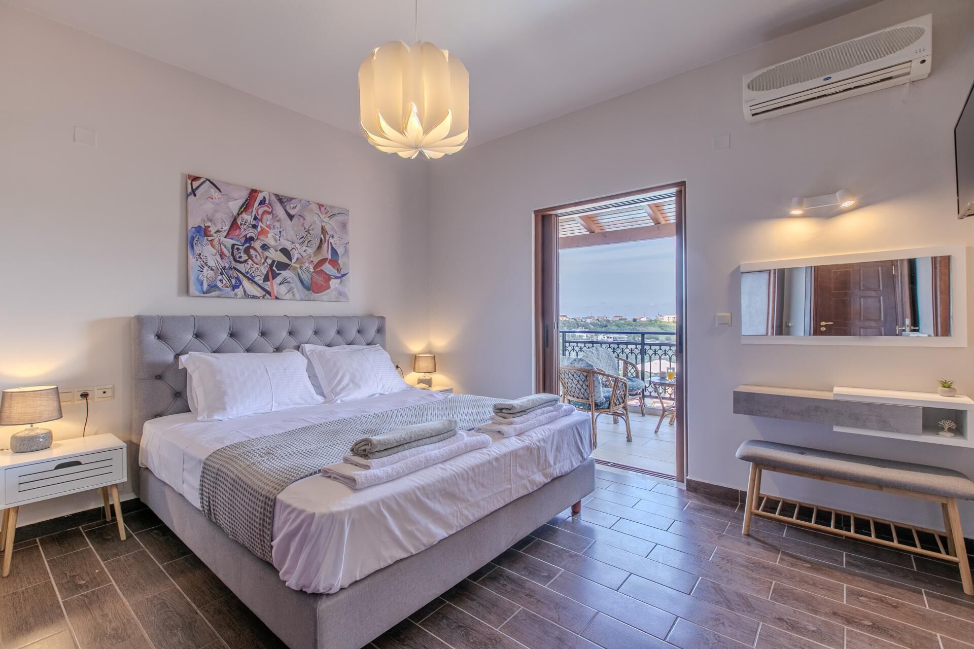 A modern bedroom with a large bed, a ceiling light, a painting, a balcony with a view, and a bathroom with a bench.