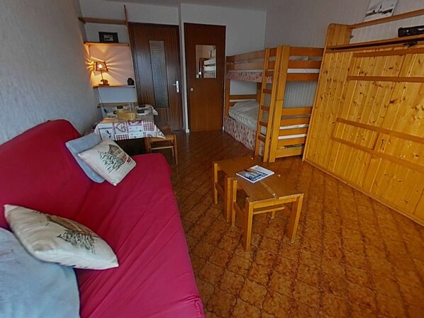 Une pièce de taille modeste, avec un canapé rouge, une petite table, un lit superposé et un bureau avec une lampe.