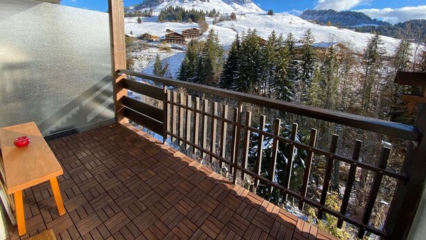Un balcón con piso de madera y una mesa, con vista a un paisaje montañoso nevado y edificios.