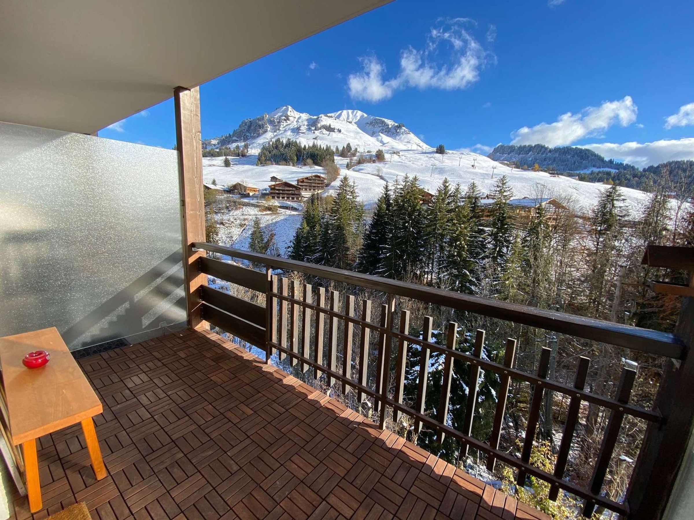 Un balcon doté d’un plancher en bois et d’une table, offrant une vue sur un paysage montagneux enneigé, avec des arbres et des bâtiments.