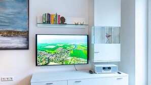 Ett modernt vardagsrum med en platt-TV som är monterad på ett vitt skåp, en bokhylla med böcker och dekorativa föremål samt en inramad tavla på väggen.