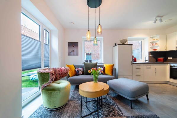 A modern living room with a sofa, ottoman, and round coffee table, a kitchen area, and large windows with blinds.