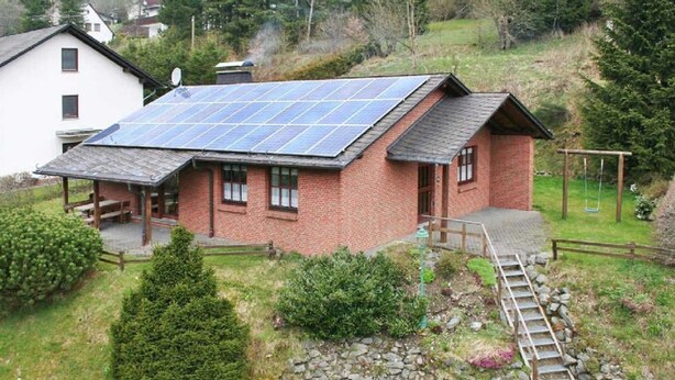Une maison avec un toit équipé de panneaux solaires, une terrasse en bois et un chemin de pierre menant à une aire de jeux avec des balançoires.