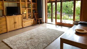 Un vaste espace de vie comprenant un téléviseur, des meubles en bois, un grand tapis et une vue sur l’extérieur grâce à une porte vitrée.
