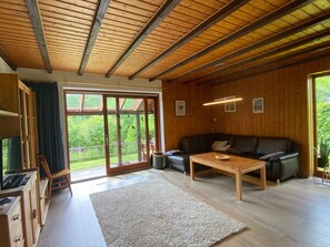 A spacious living area with a wooden ceiling, a black leather sofa, a wooden coffee table, and a large sliding glass door leading to a balcony.