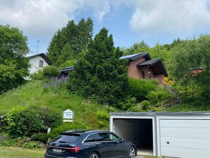 En bil parkeret foran en bygning med garage, omgivet af grøn vegetation og træer.