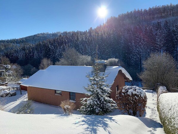 Une maison recouverte de neige, avec un ciel dégagé et une colline boisée en arrière-plan.