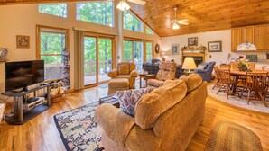 A spacious living room with a large sofa, a television, and a fireplace. There are multiple windows providing natural light and a view of the outdoors.