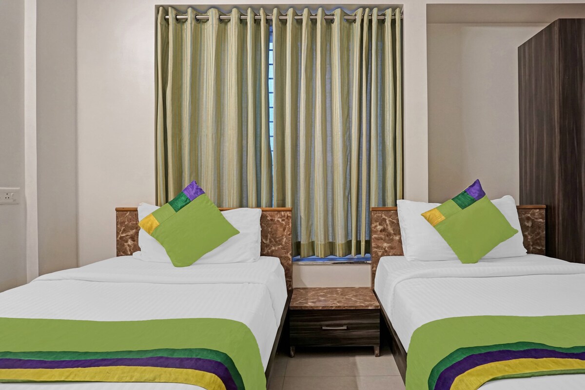 A hotel room with two beds, a small wooden nightstand, and a window with vertical blinds.
