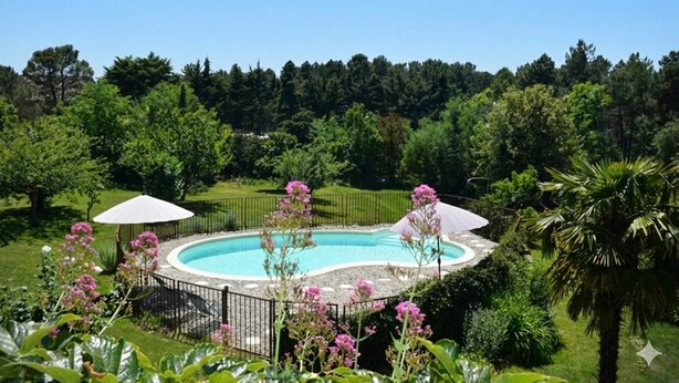 A circular pool surrounded by a black fence, with two white umbrellas and pink flowers in the foreground. - GÎte Les Cerisiers, 3 stars (Ailhon)