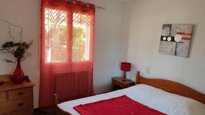 A bedroom with a wooden bed, a red throw blanket, a nightstand with a lamp, a dresser, a window with sheer curtains, and a wall-mounted abstract painting. - GÎte Les Cerisiers, 3 stars (Ailhon)
