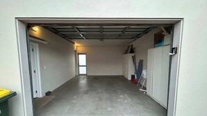 A garage with a concrete floor, a white door, and a metal garage door. - Central Oasis - Garage & Courtyard, Sleeps 6 (Rotorua)