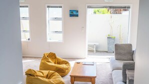 A spacious living room with a grey sofa, yellow bean bag chairs, a wooden coffee table, and a large sliding glass door leading to an outdoor area. - Central Oasis - Garage & Courtyard, Sleeps 6 (Rotorua)