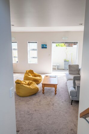 Ein geräumiges Wohnzimmer mit grauem Sofa, gelben Sitzsäcken, einem Holztisch und einer großen Schiebeglastür, die zu einem Außenbereich führt.