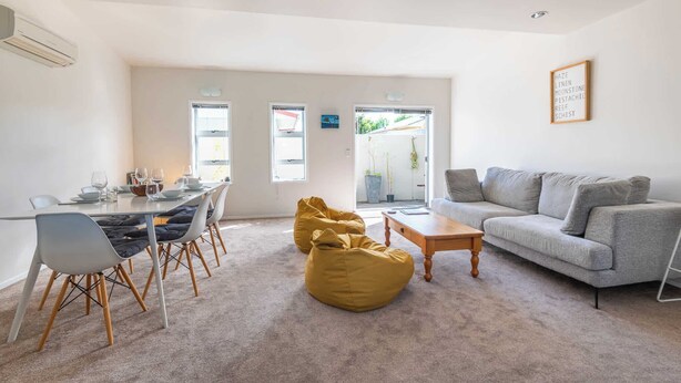 A modern living room with a grey sofa, a wooden coffee table, and a yellow bean bag. There is a dining area with a table set for four, and a large window providing natural light. - Central Oasis - Garage & Courtyard, Sleeps 6 (Rotorua)