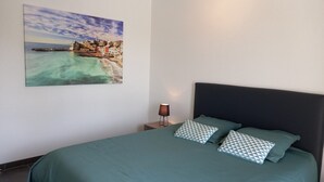 A bedroom with a bed, pillows, a nightstand, a lamp, and a painting of a coastal town. - Sarlajoande, a modern villa in Perigord style (Proissans)