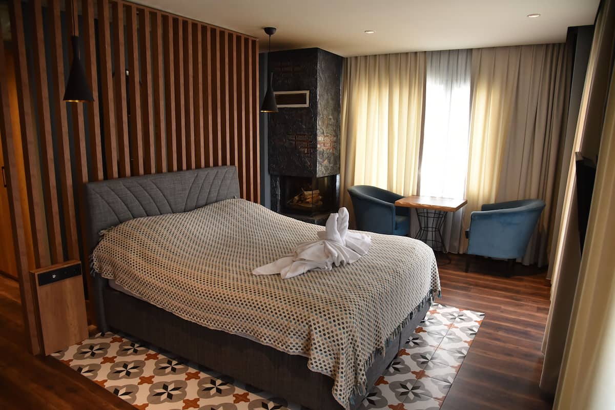 A modern hotel room with a large bed, a fireplace, a dining area, and a view of the outdoors.