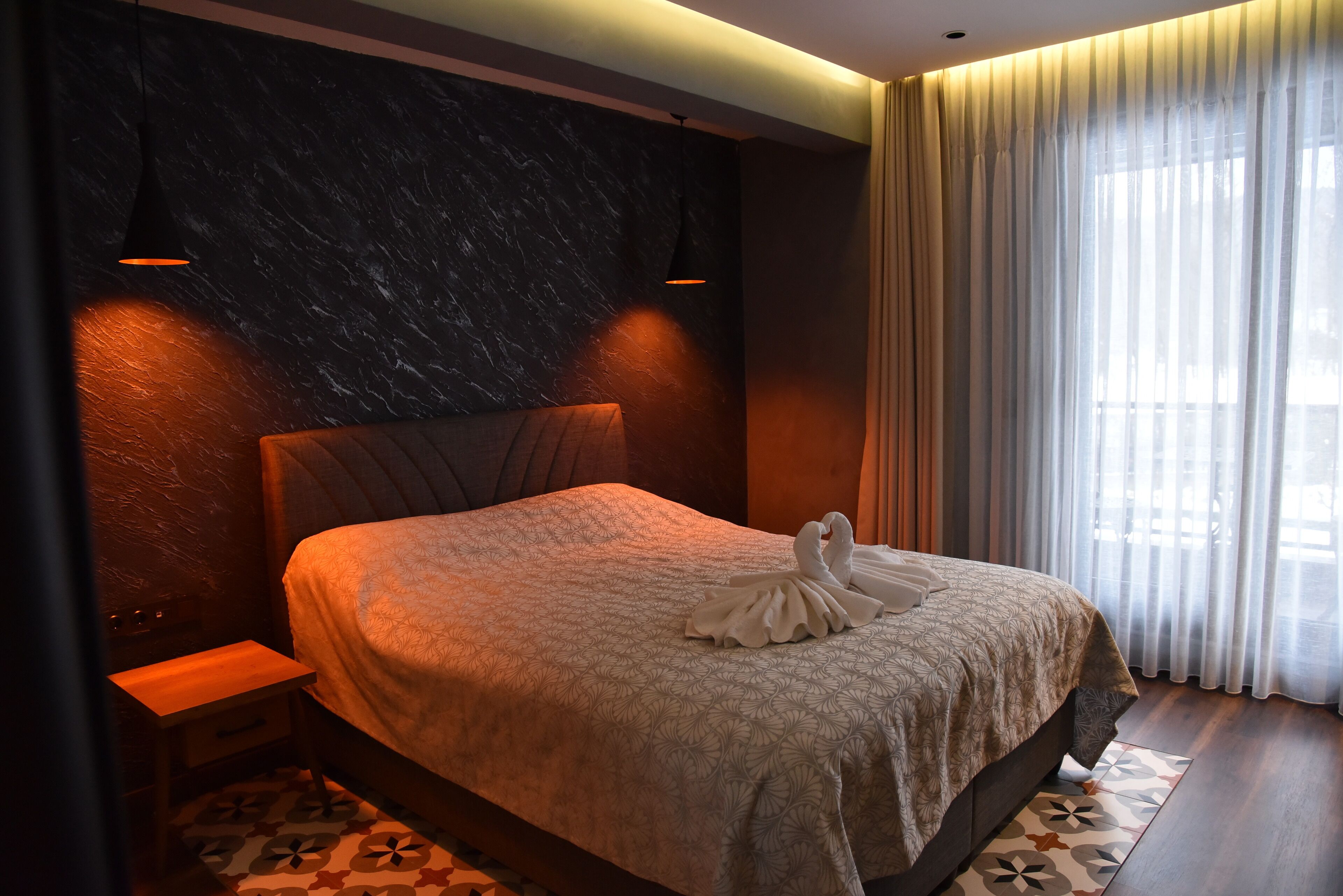 Una cama impecable con una toalla con forma de cisne, una pared de mármol oscuro y una ventana con cortinas traslúcidas.