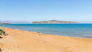 Une plage de sable fin, bordée d’une eau d’un bleu limpide et dominée par des collines rocheuses à l’horizon.