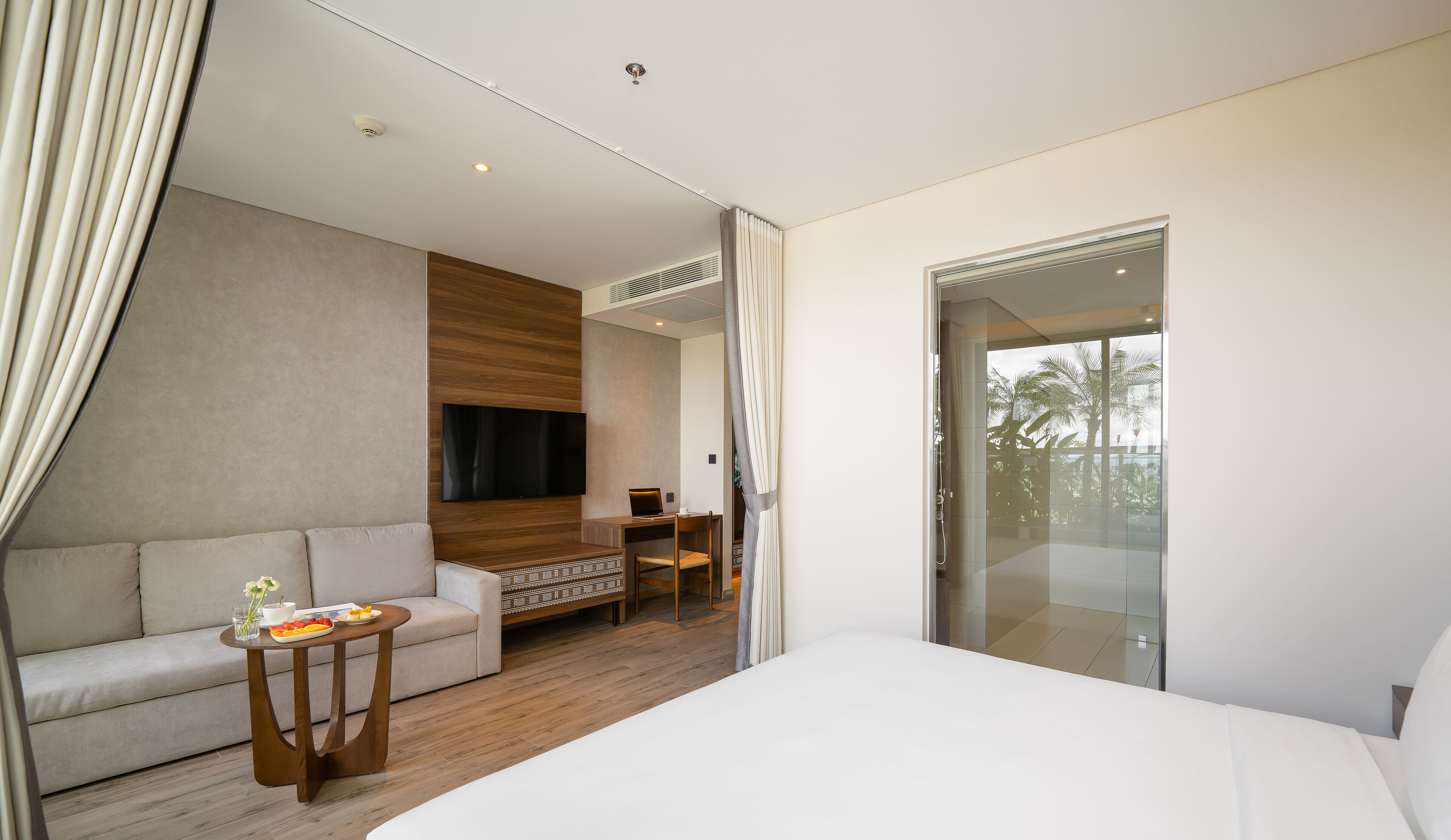 Ein modernes Hotelzimmer mit einem großen Bett, einer Couch, einem kleinen Tisch mit Obstteller, einem Schreibtisch mit Laptop, einem an der Wand montierten Fernseher und einer Glastür zum Balkon mit Blick auf Palmen.